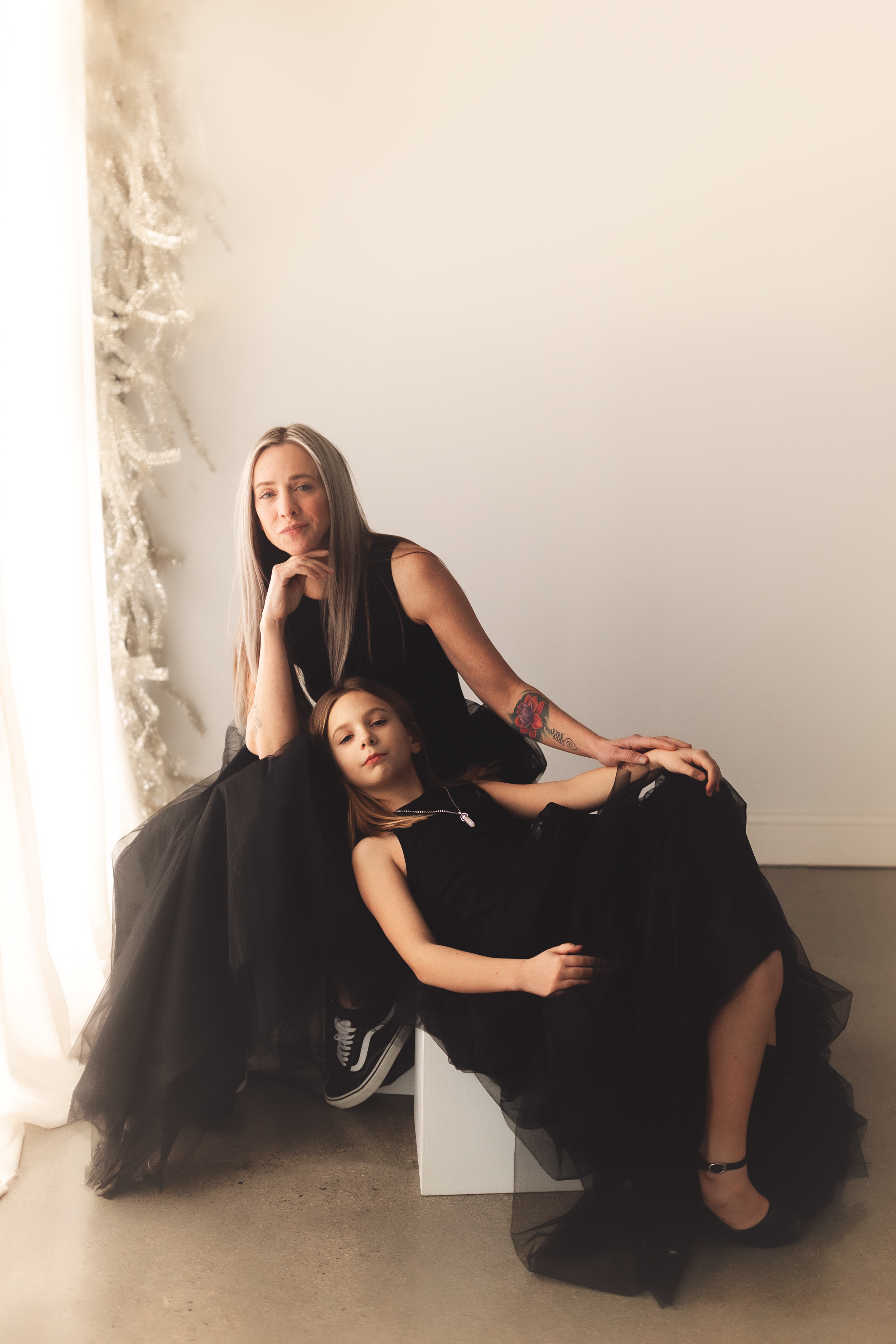 Two people in black dresses pose dramatically in an artistic photoshoot against a white wall.