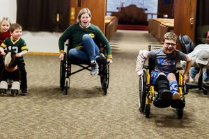 Young students in wheelchairs practice using adaptive equipment in a school hallway.