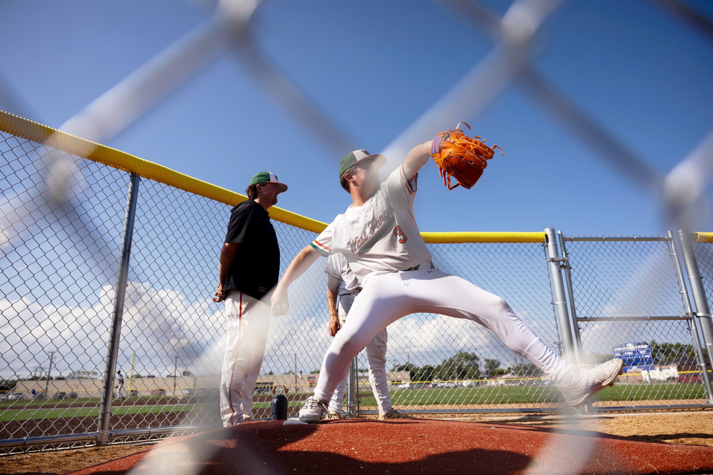 A pitcher in white uniform winds up to throw during a baseball game, viewed through the mesh of a chain-link fence.