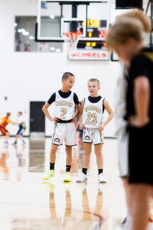 Two young basketball players in white jerseys stand together on a court during a game or practice session.