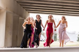 Wedding party in formal dresses runs playfully under a bridge, showing candid joy and movement.