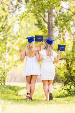 Two graduates in white dresses walk together on a sunny park path holding blue diplomas behind their backs.
