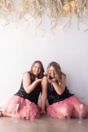 Two friends wearing matching black sequin tops and pink tulle skirts laugh together under sparkly decor.
