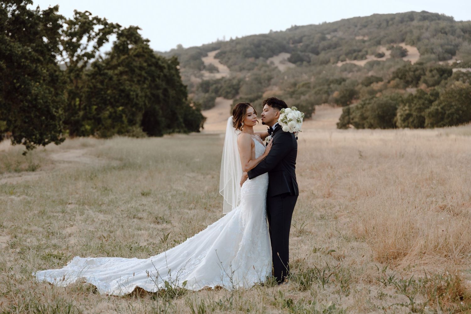 A wedding couple embraces in a golden field with rolling hills in the background at sunset.