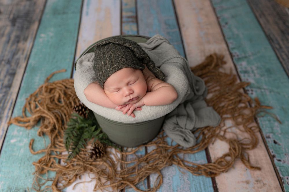 Sleeping newborn in grey wrap positioned in bowl on rustic turquoise wooden backdrop.