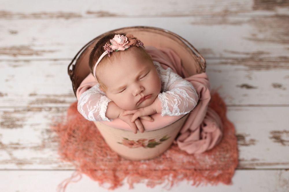 Sleeping baby in vintage bowl with lace outfit and headband on rustic white wooden background.