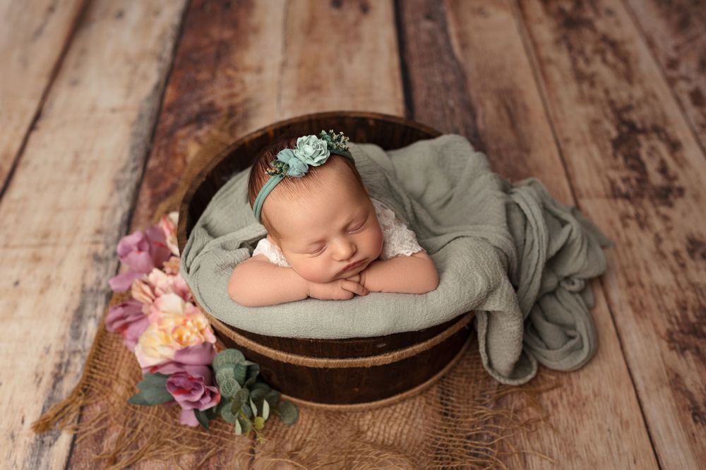 Peaceful newborn sleeping in round basket with flowers and gray fabric on wooden background.