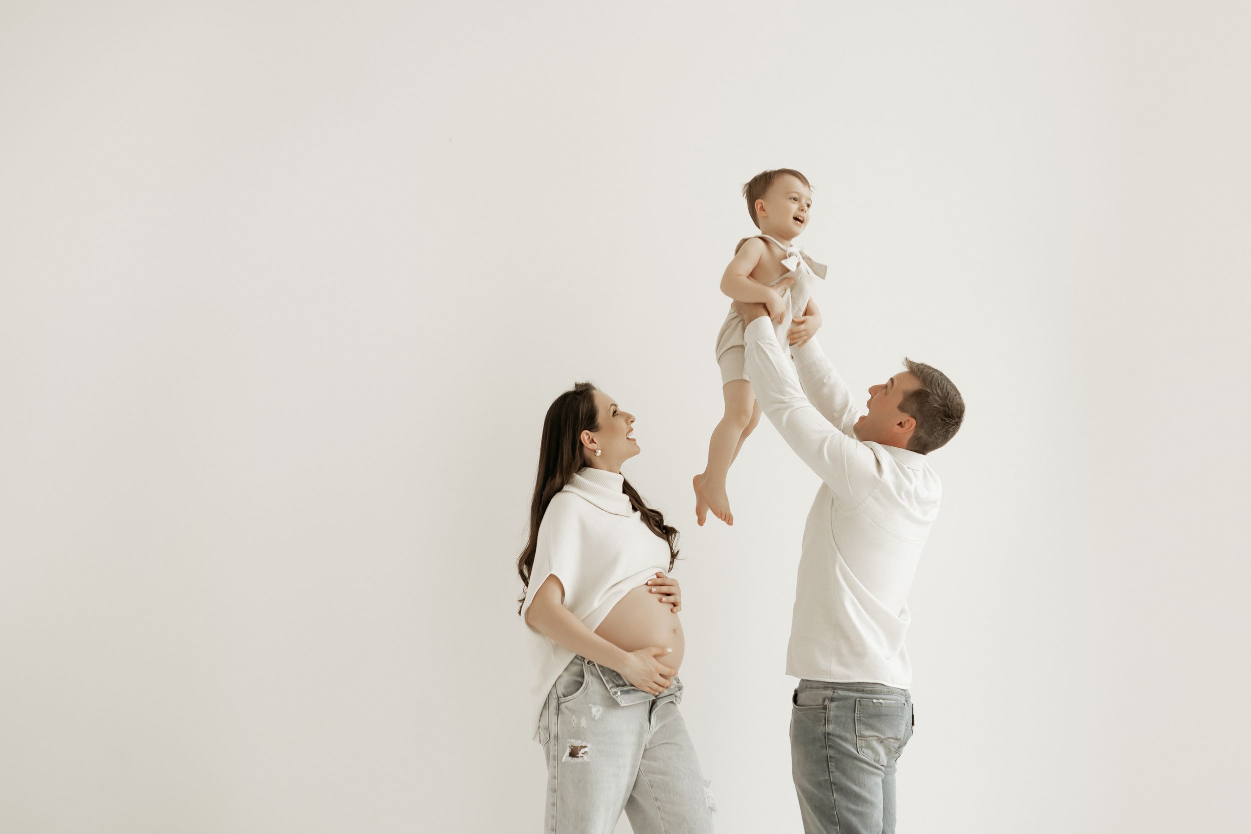 Family portrait of parents lifting baby in the air against white wall.