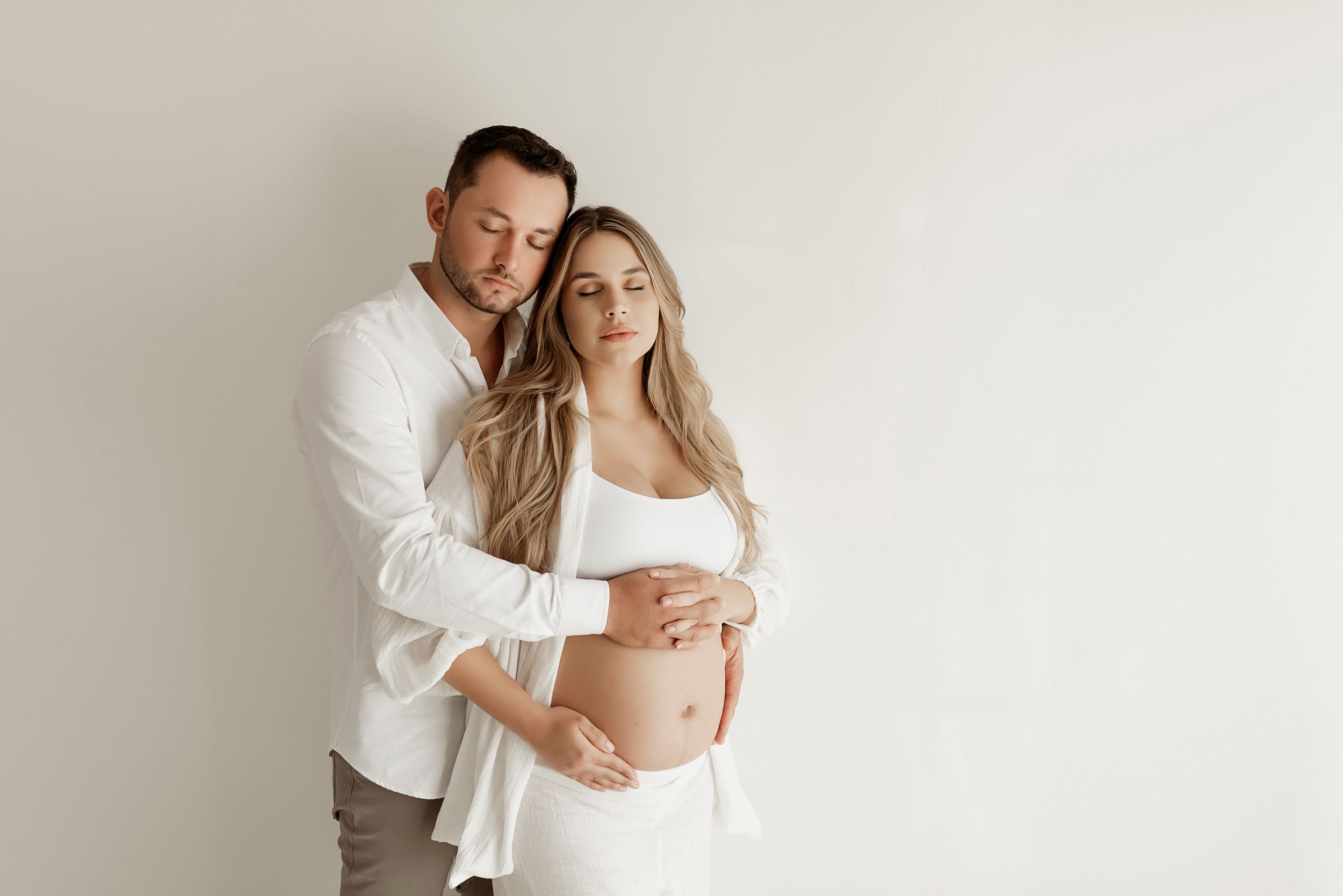 Intimate maternity portrait of couple in white clothing against neutral background.