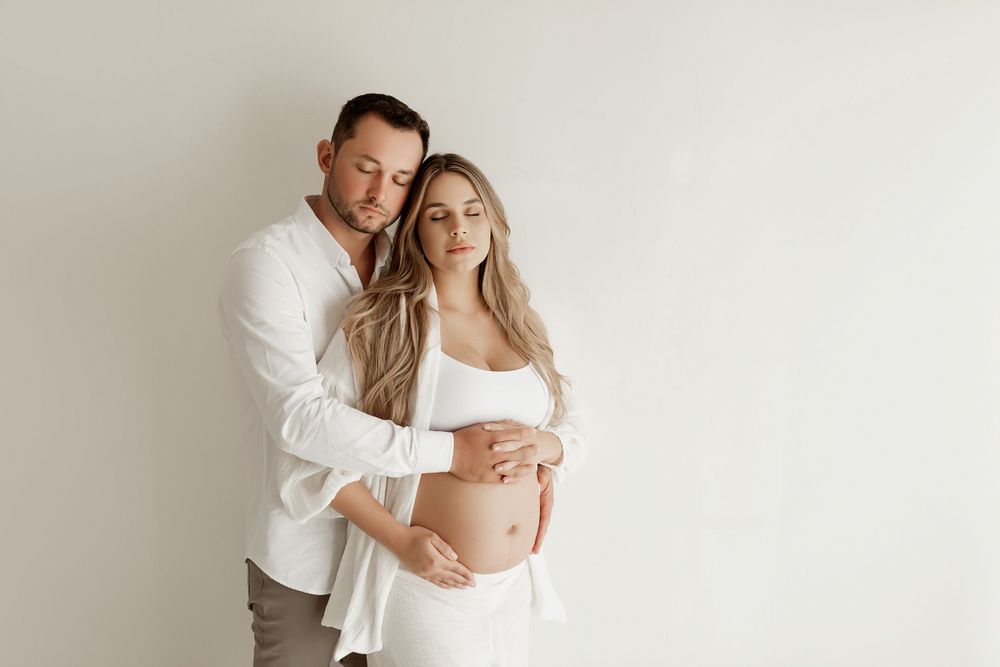 Intimate maternity portrait of couple in white clothing against neutral background.