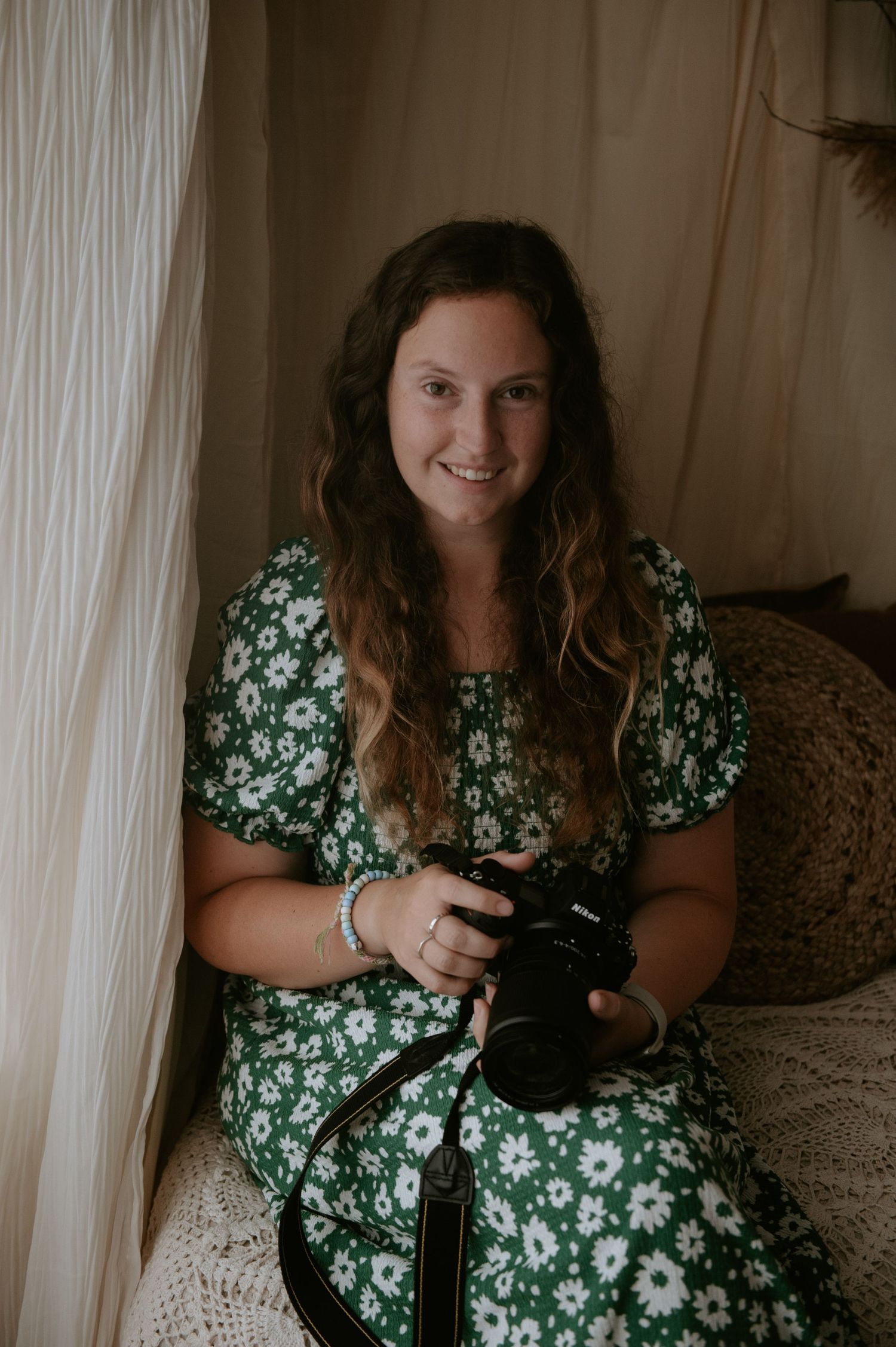 Photographer in a green floral dress sits by a window holding a professional camera.