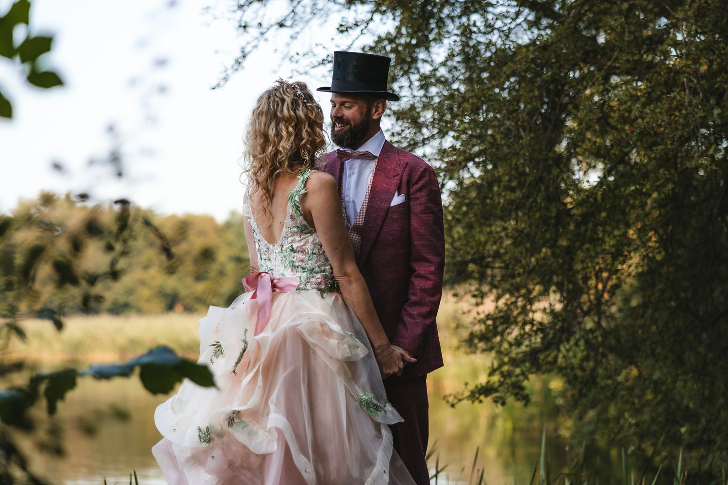 A couple in elegant wedding attire share a romantic moment under trees by a pond on their special day.