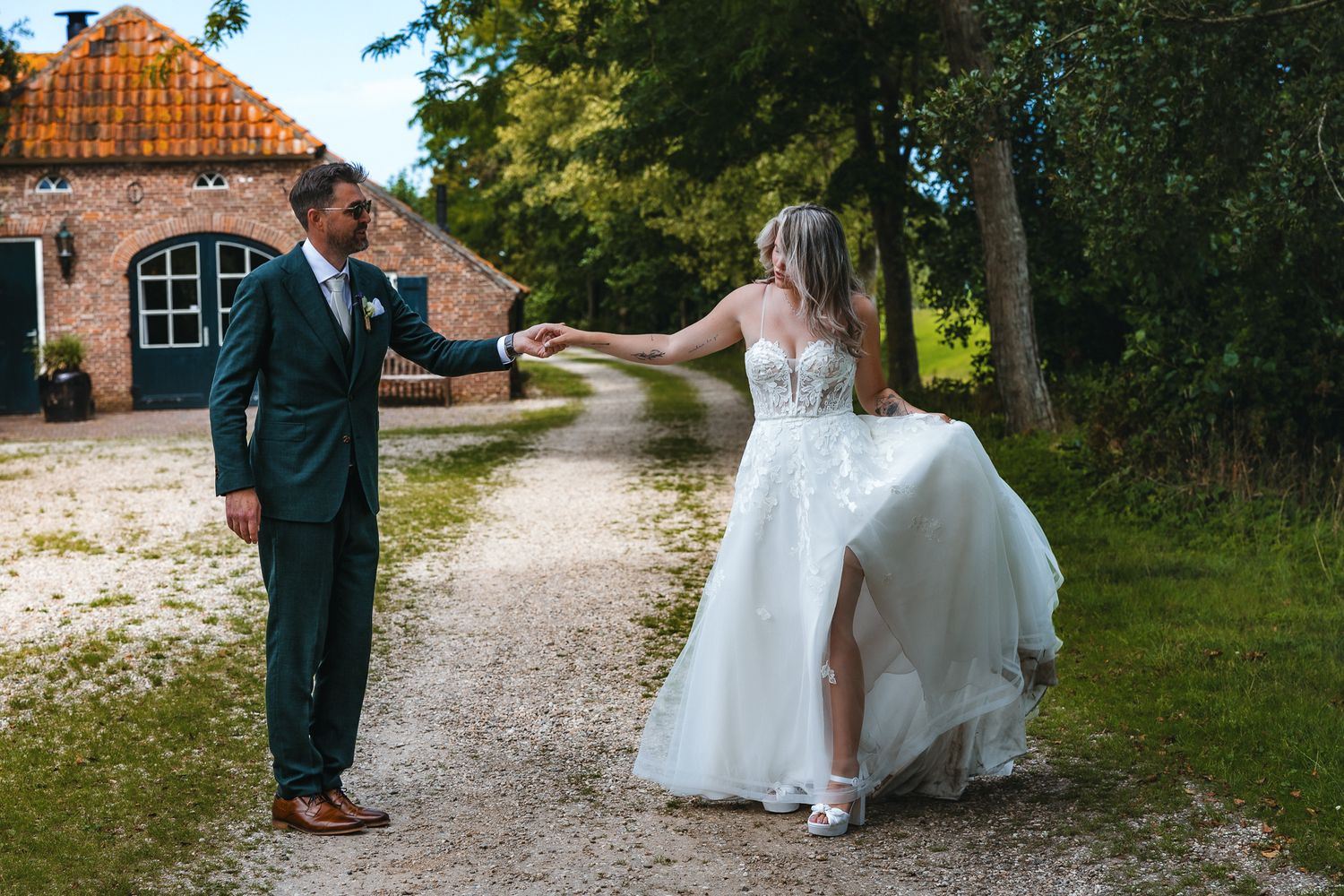 A couple in wedding attire dances joyfully on a rustic gravel path near an old brick building on their special day.