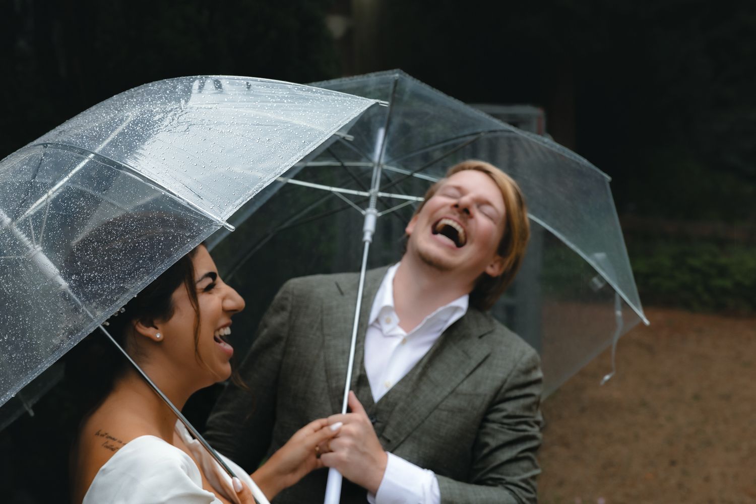 Two people share an umbrella and laugh joyfully in the rain while wearing formal attire.