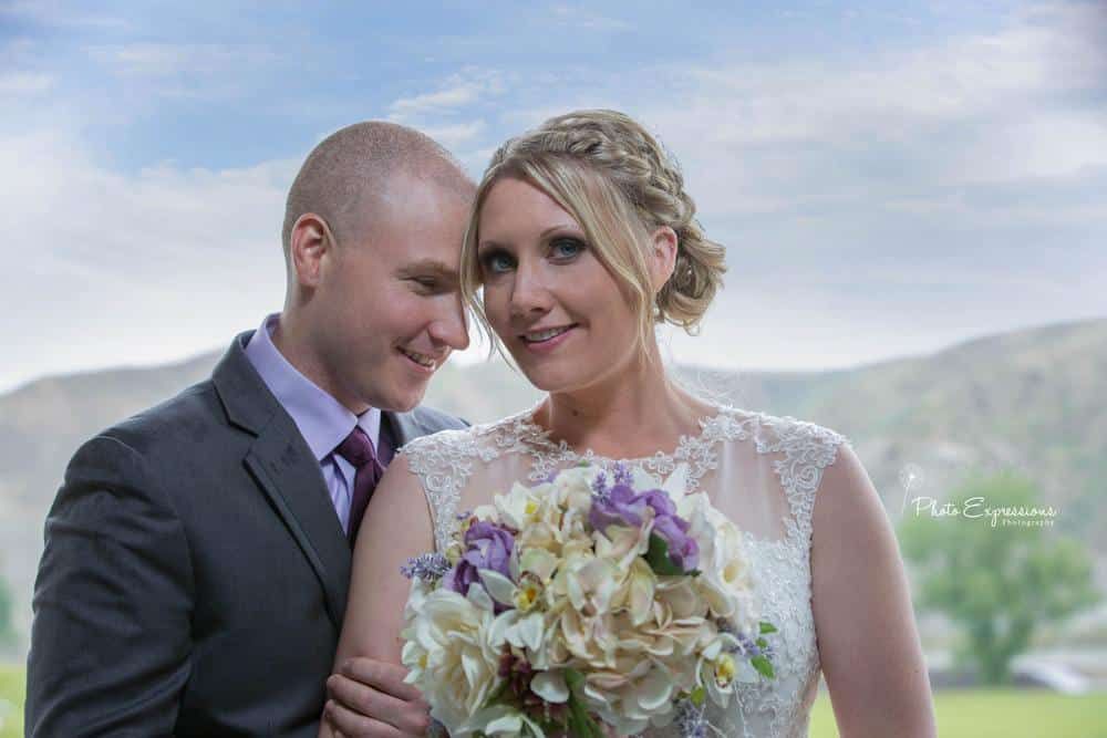 A bride and groom embrace on a grassy hillside with mountains in the background.