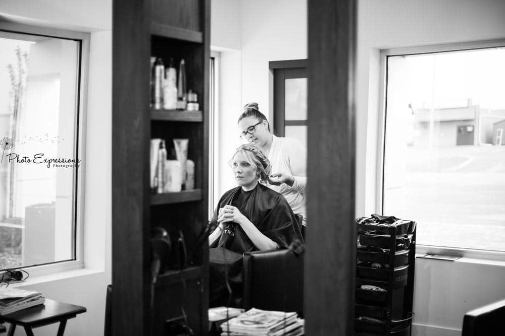 Black and white photo showing preparation in a salon with styling chair and shelves visible in background.