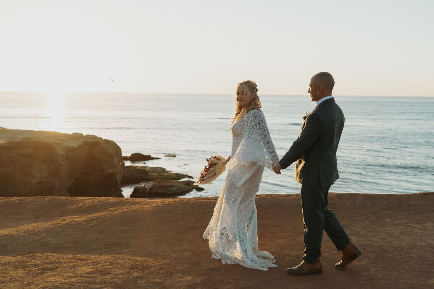 A couple walks along a rocky beach at sunset in formal wedding attire holding hands.