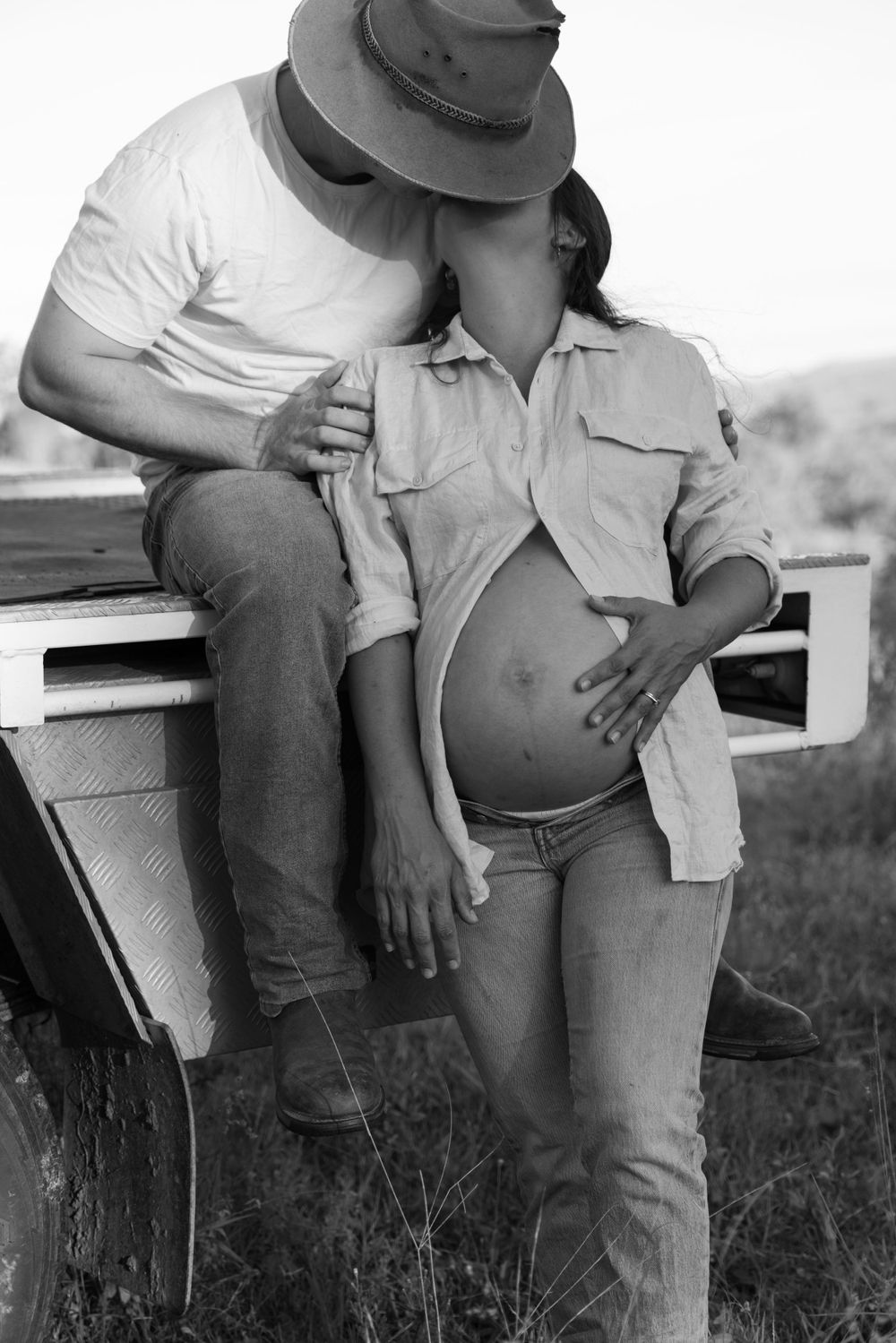 Expecting parents casually leaning against ute in outdoor countryside setting, kissing.