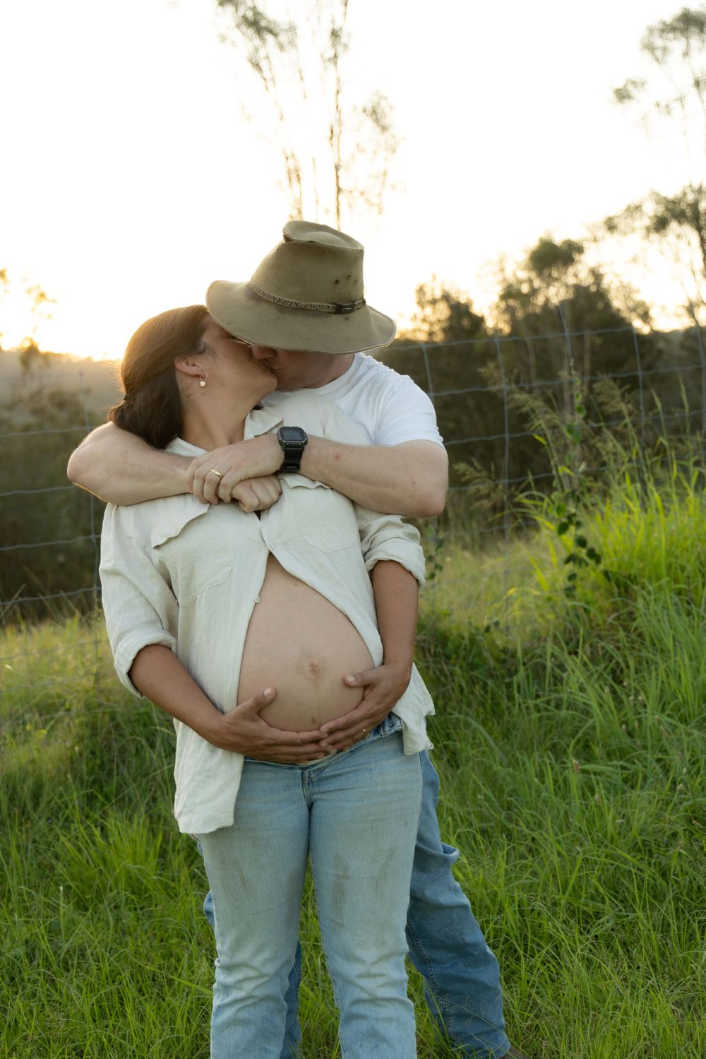 A sunset maternity photoshoot shows a tender embrace between expecting parents in a grassy field.