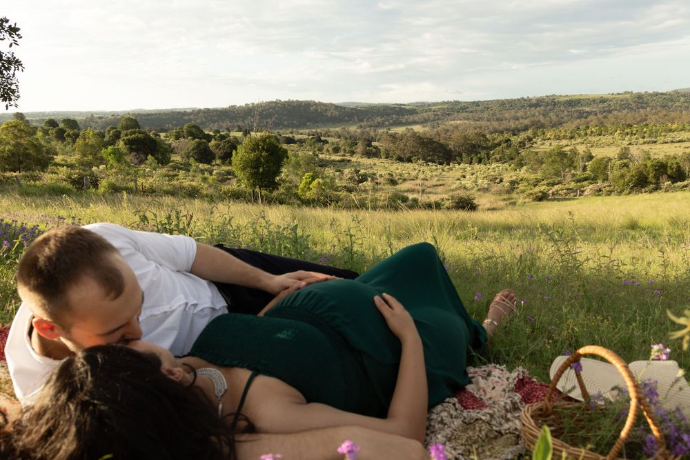 Rolling hills and grassland landscape stretches into the distance under a cloudy sky. Expecting couple laying on mat.
