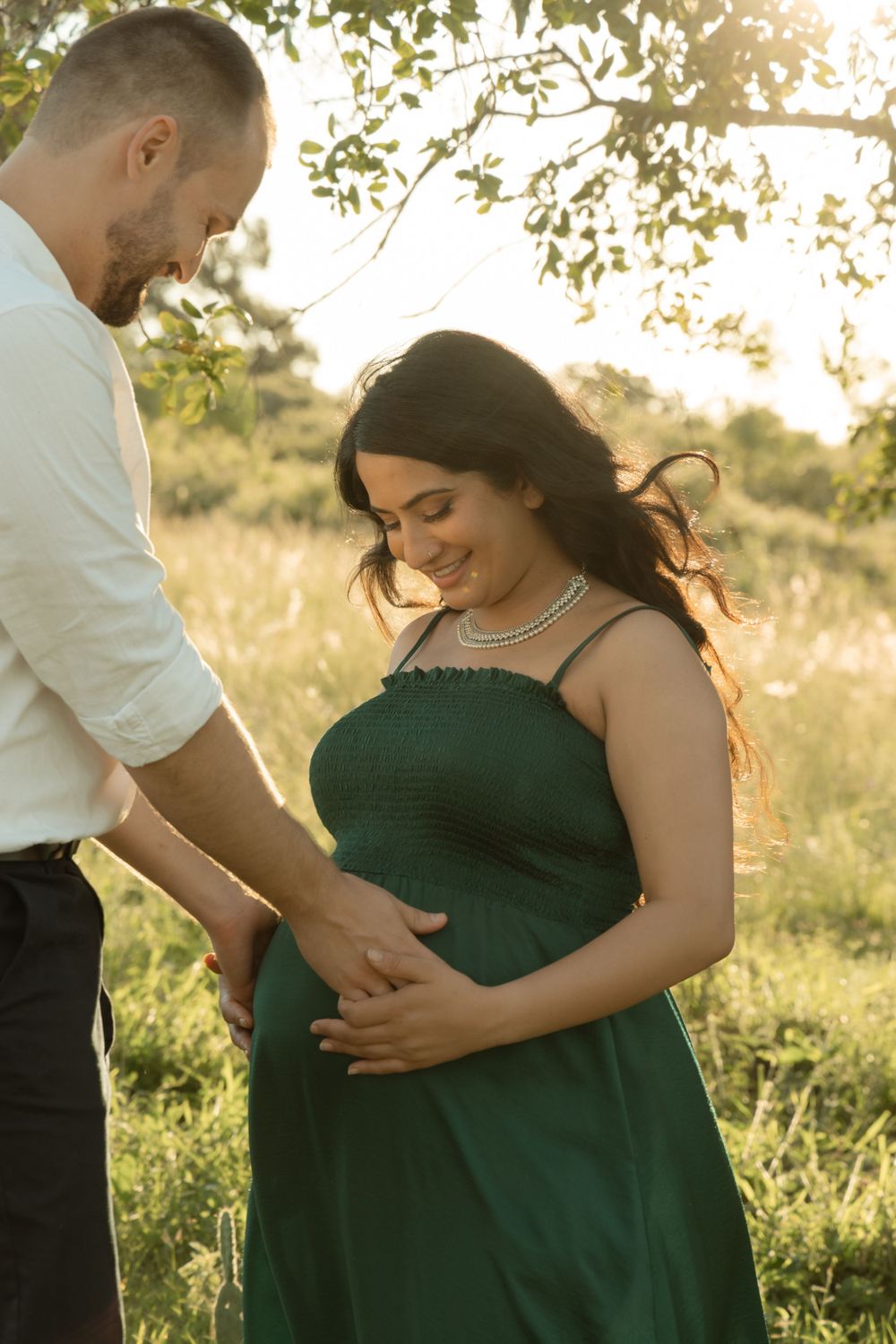 A couple in a field with pregnant woman wearing green dress during golden hour photography session.