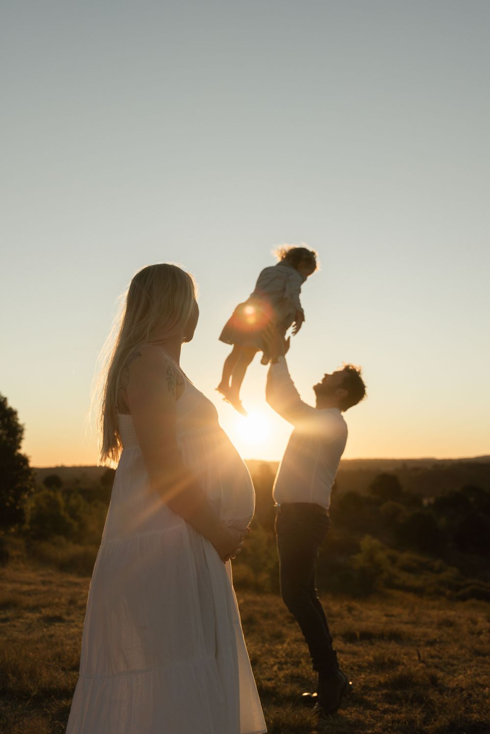 A couple shares tender moments during their sunset maternity photoshoot. Father throwing daughter in air. Mother holding her bump.