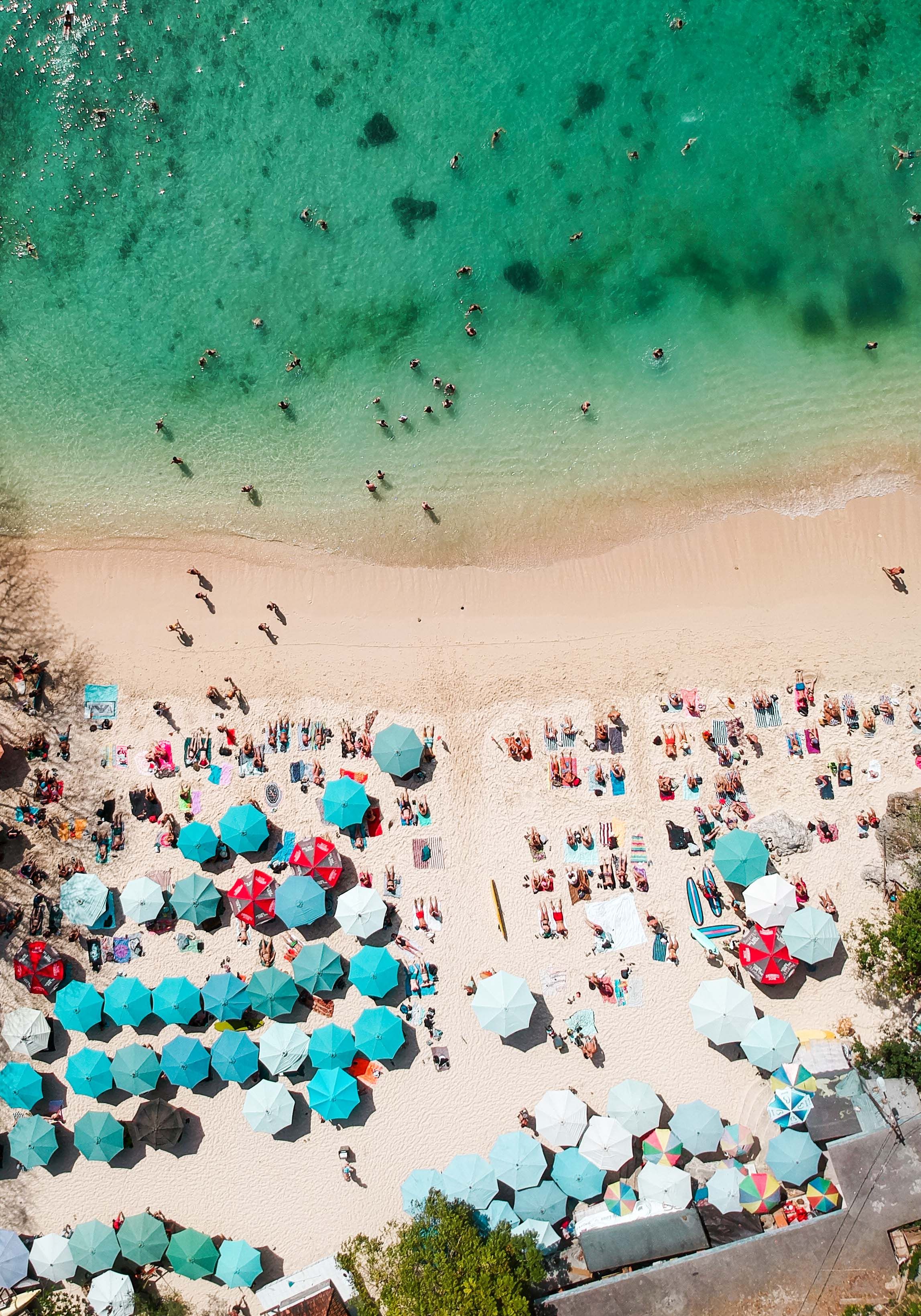 Aerial view of a crowded beach with turquoise water and bright blue umbrellas dotting the sandy shore.