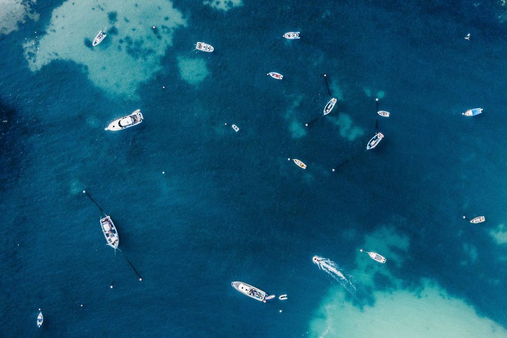 Boats and yachts scattered across bright blue tropical waters viewed from above.