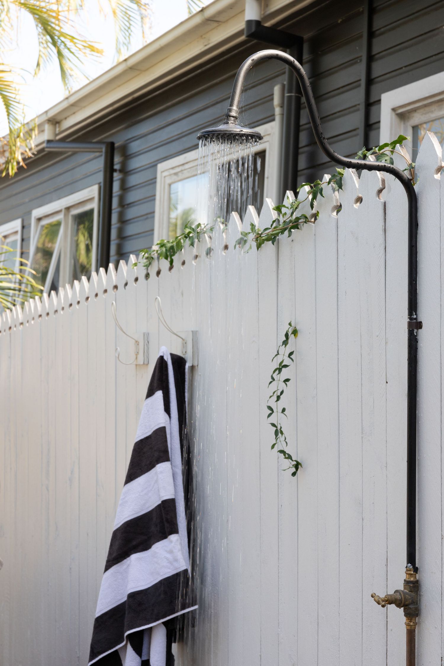 Outdoor shower with striped towel and climbing vines against white fence.