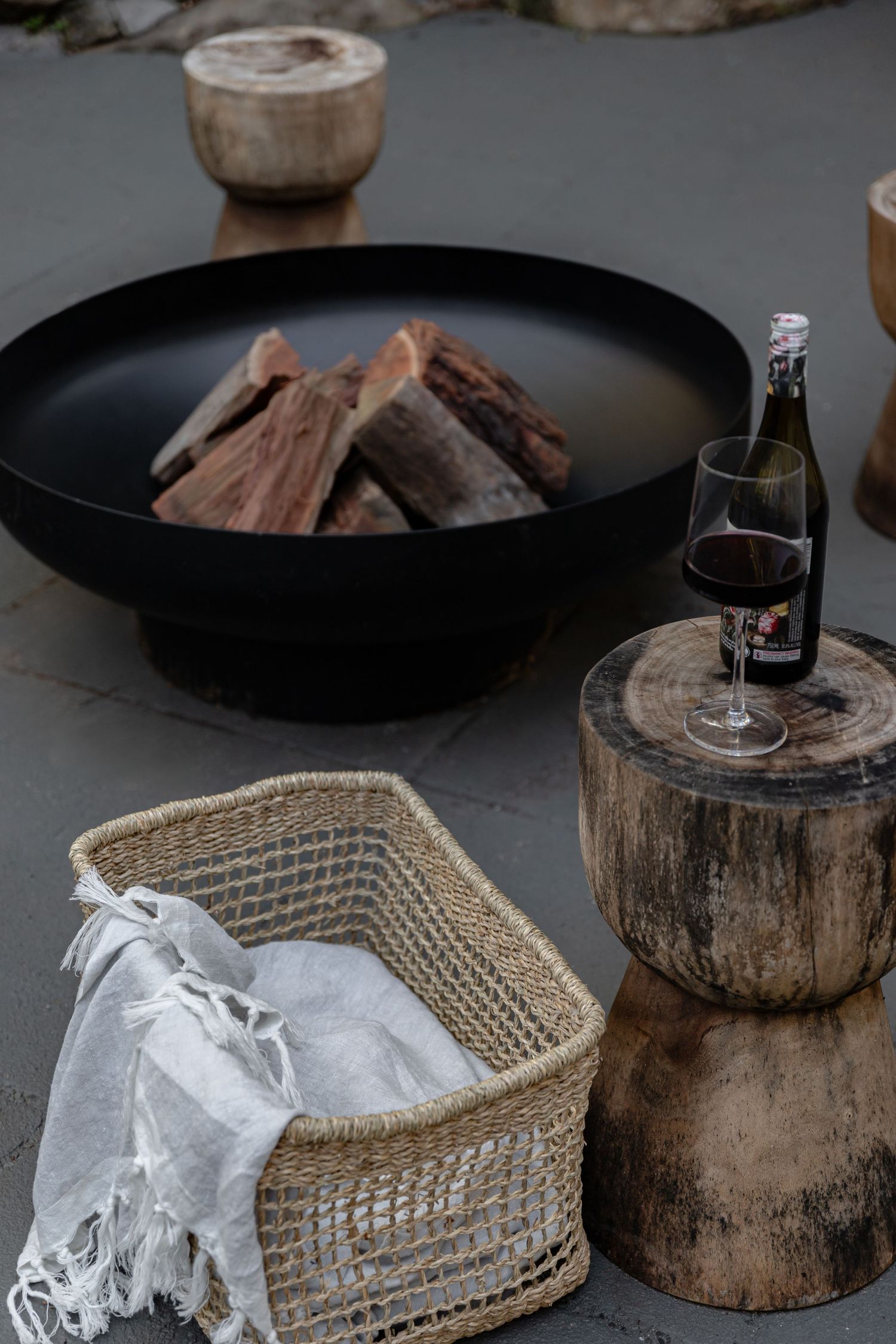 Rustic outdoor scene with fire pit, wine glass on wood stump, and woven basket with white textile.