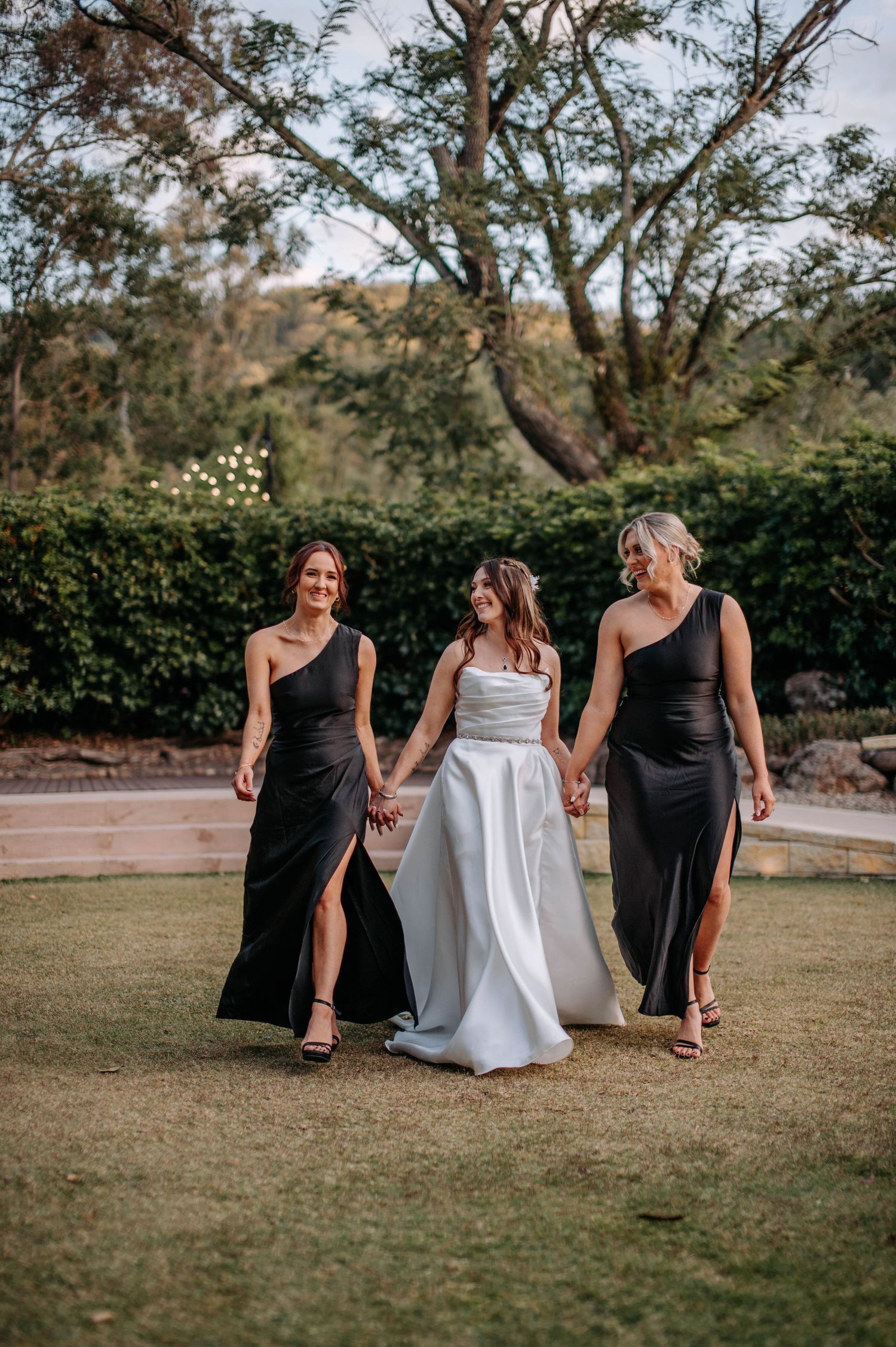 Three elegantly dressed women walk together on garden lawn during wedding celebration.