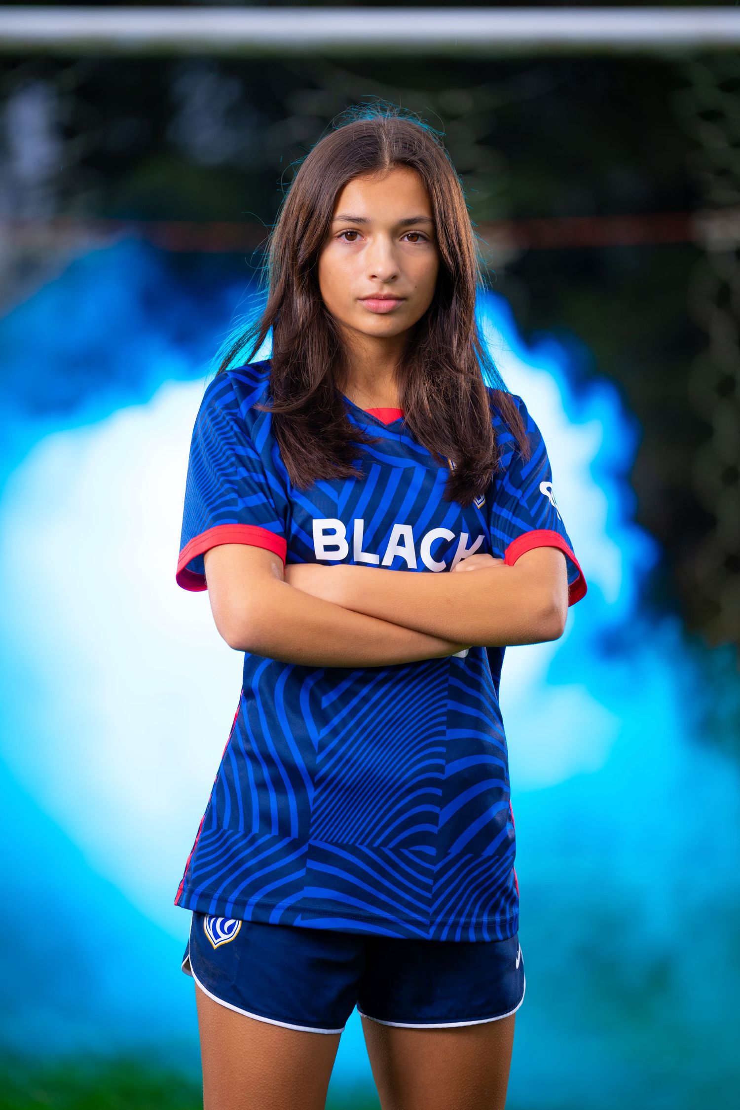 A female soccer player from Reign Academy poses against a backdrop of a soccer goal and smoke, lit by a blue light.