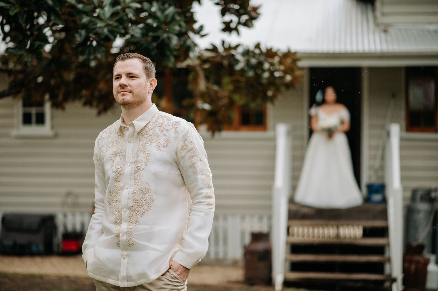 A groom in traditional white Barong Tagalog stands in front of a house on his wedding day.