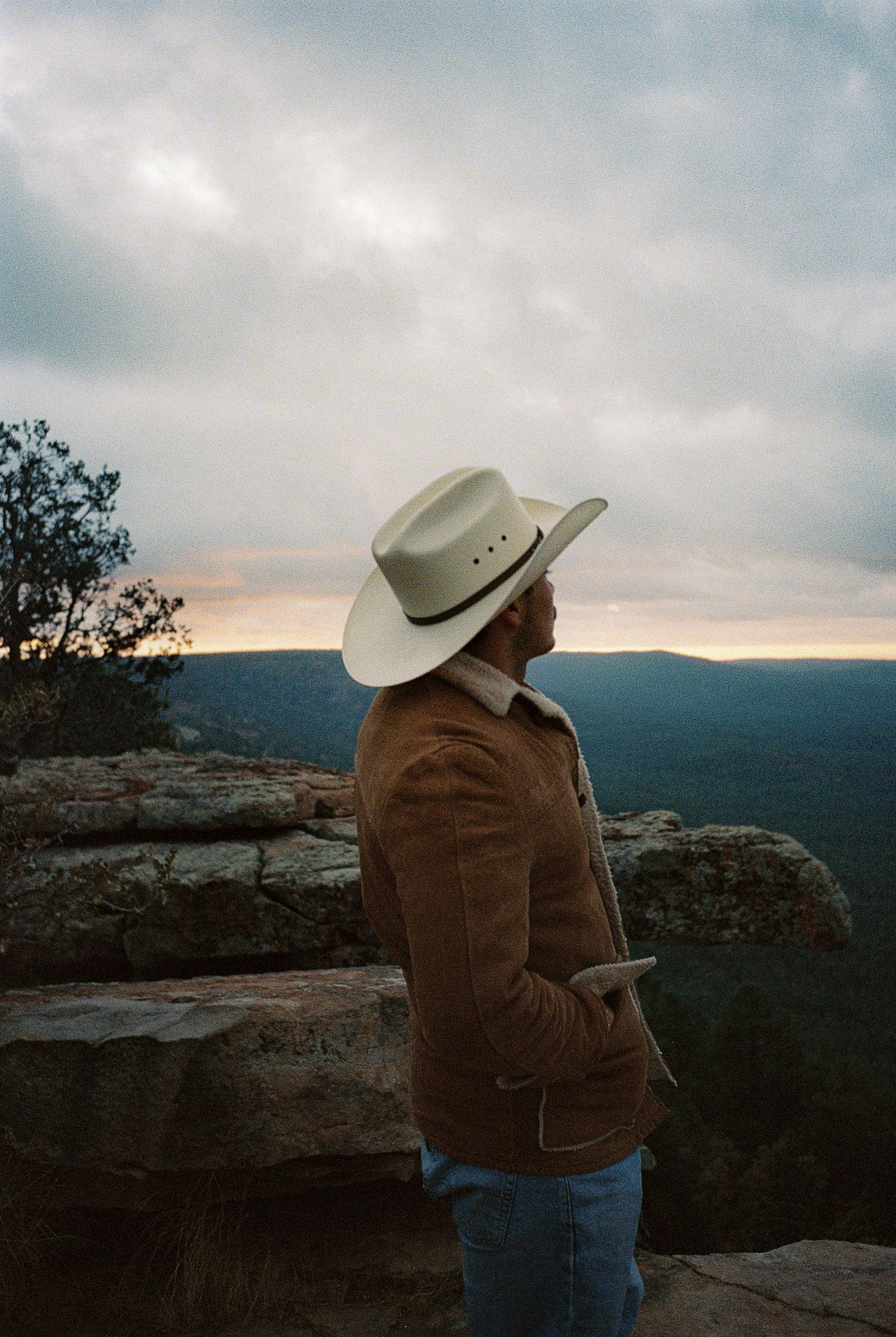 Person in cowboy hat and brown jacket overlooking a mountainous landscape at sunset.