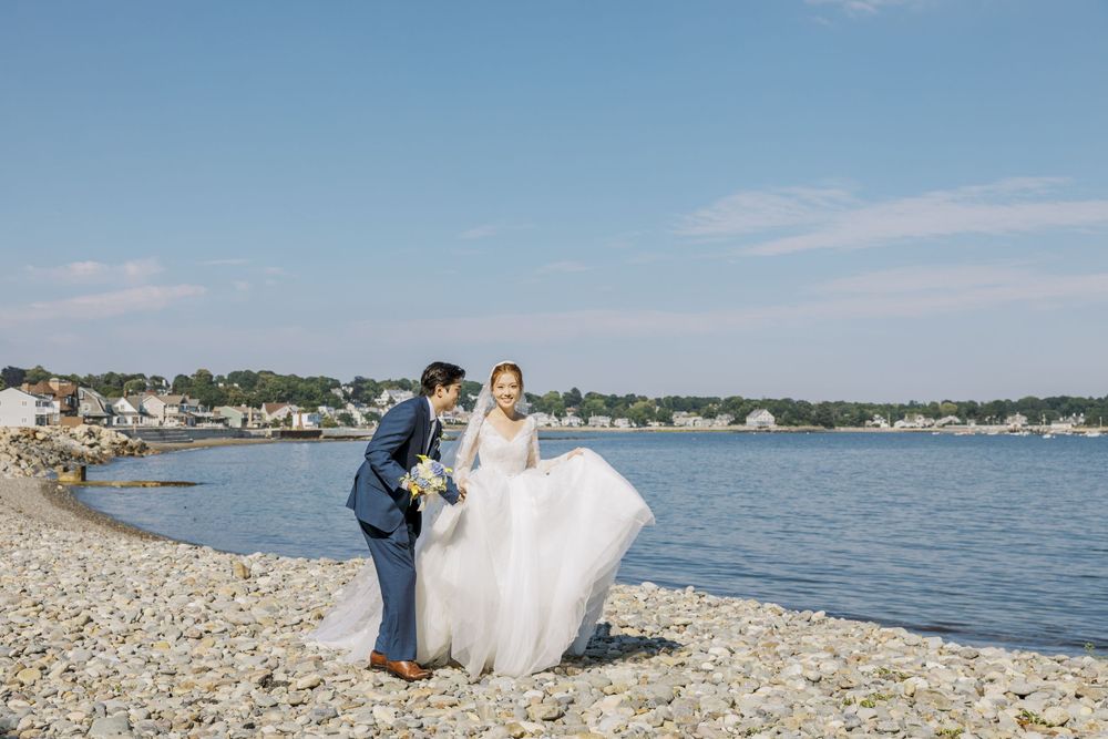 Coastal wedding photo on rocky beach with waterfront view and blue skies.