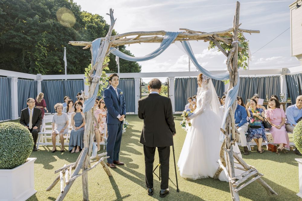 Wedding ceremony under rustic arch with blue draping and guests seated on lawn.