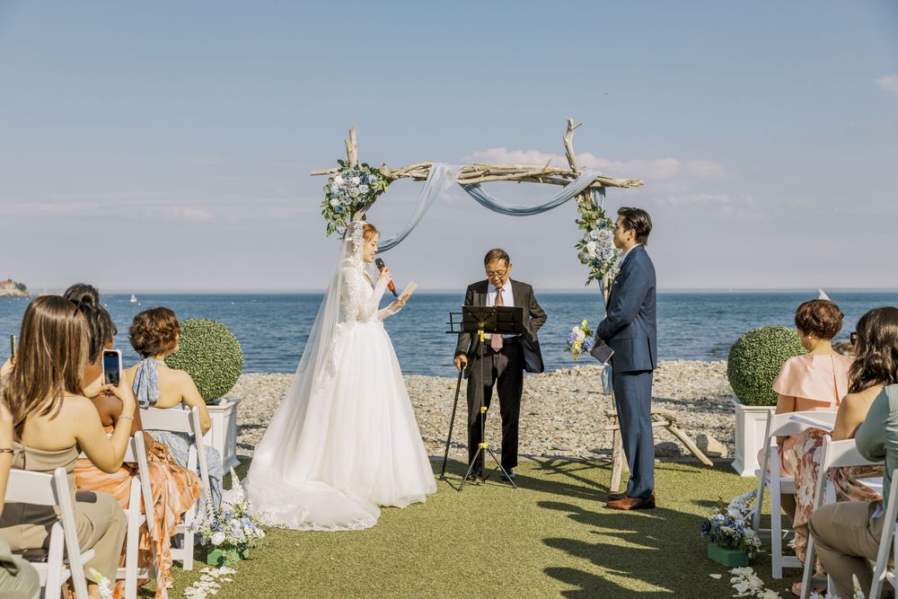 Waterfront wedding ceremony with guests seated on white chairs and rustic floral arch.