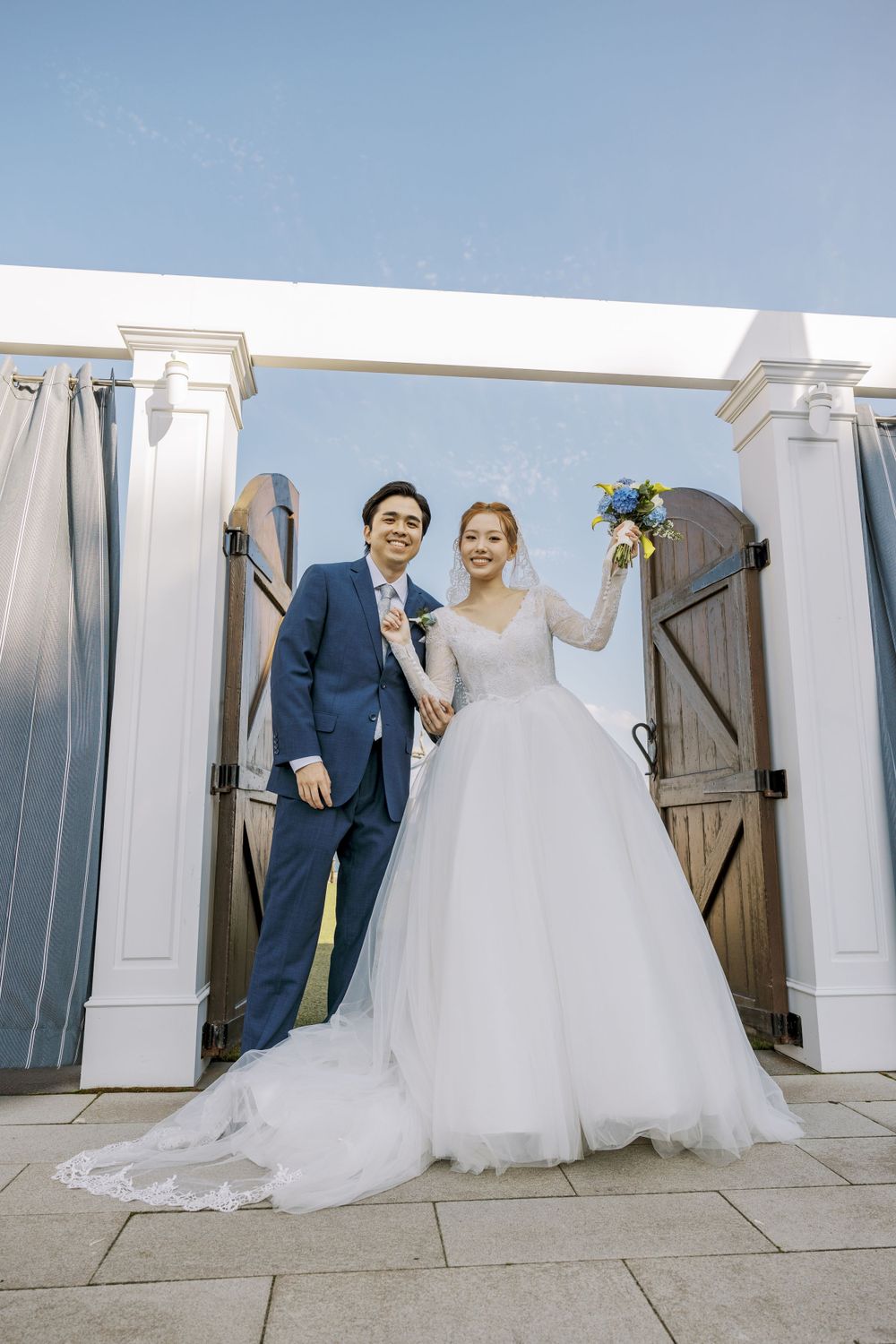 Wedding couple poses playfully behind white doors in an outdoor setting.