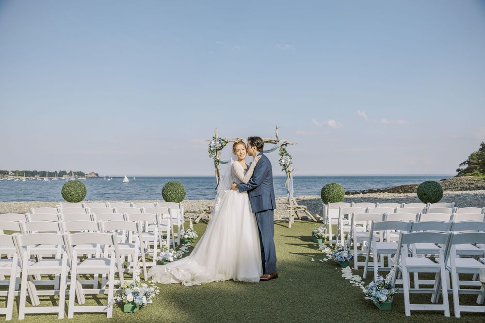 Oceanside wedding ceremony setup with white chairs and floral decorations against a blue sky.