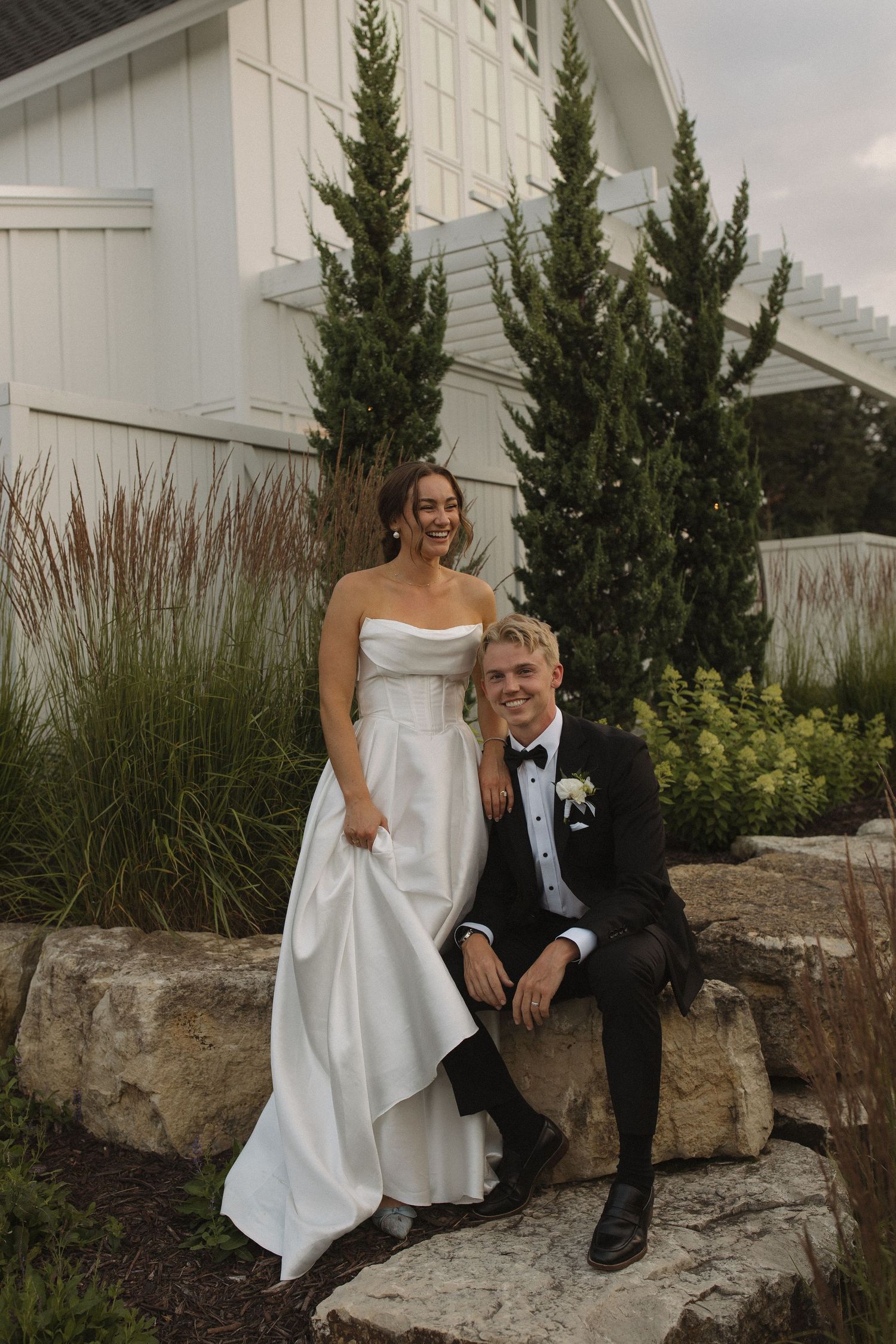 Wedding couple sitting on stone wall by white building with evergreen shrubs.