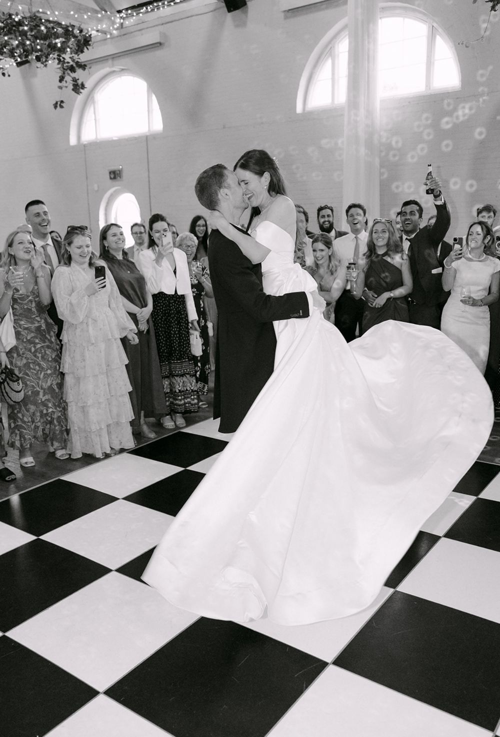 A bride twirls her dress on a black and white checkered dance floor while wedding guests watch and cheer from the sides.