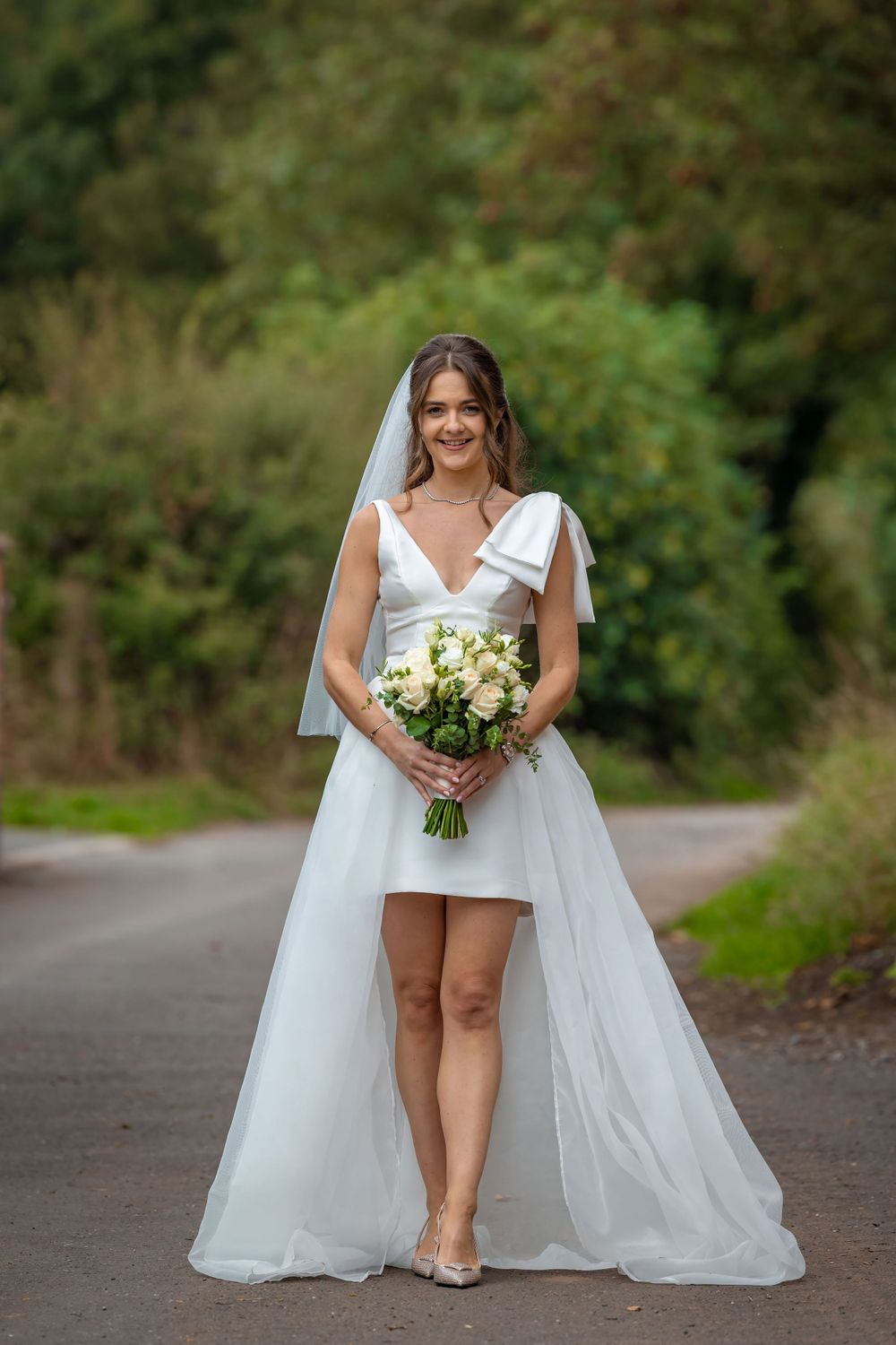Bride in a short white wedding dress and veil holding a bouquet on a garden path.