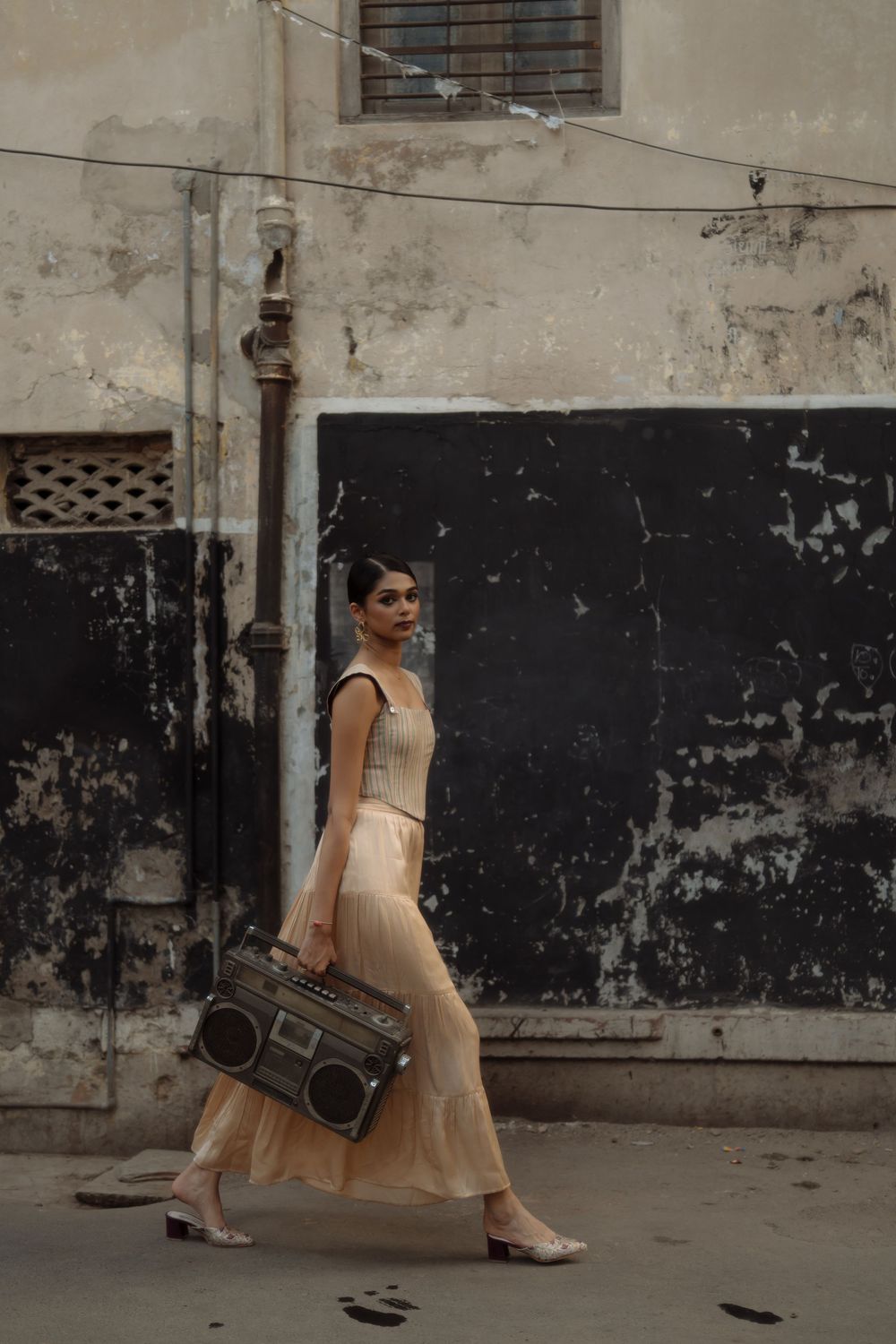 Dancing figure in flowing beige dress poses with vintage boombox against weathered urban wall.