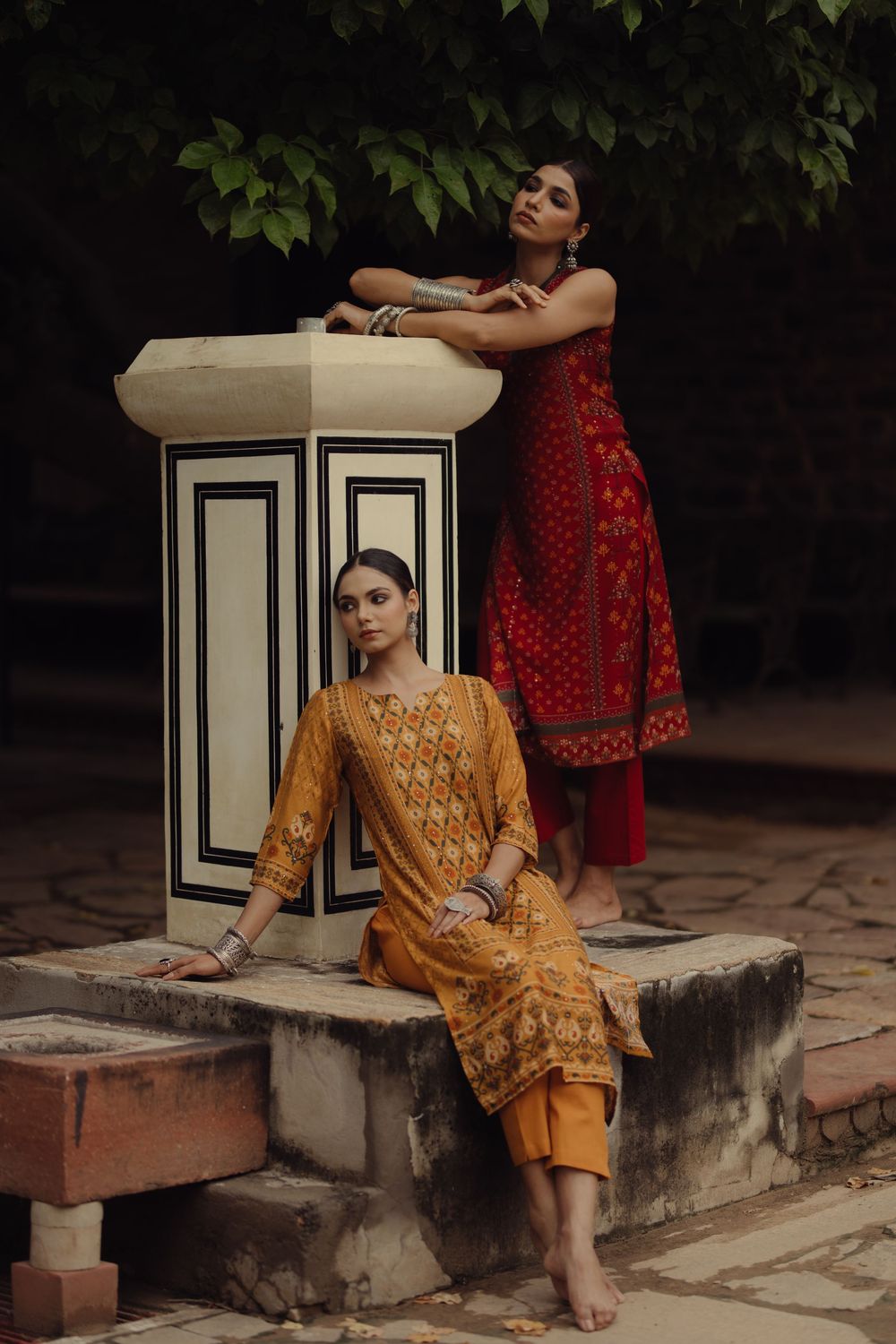 Two women in traditional South Asian fashion pose elegantly against a white pillar and garden backdrop.