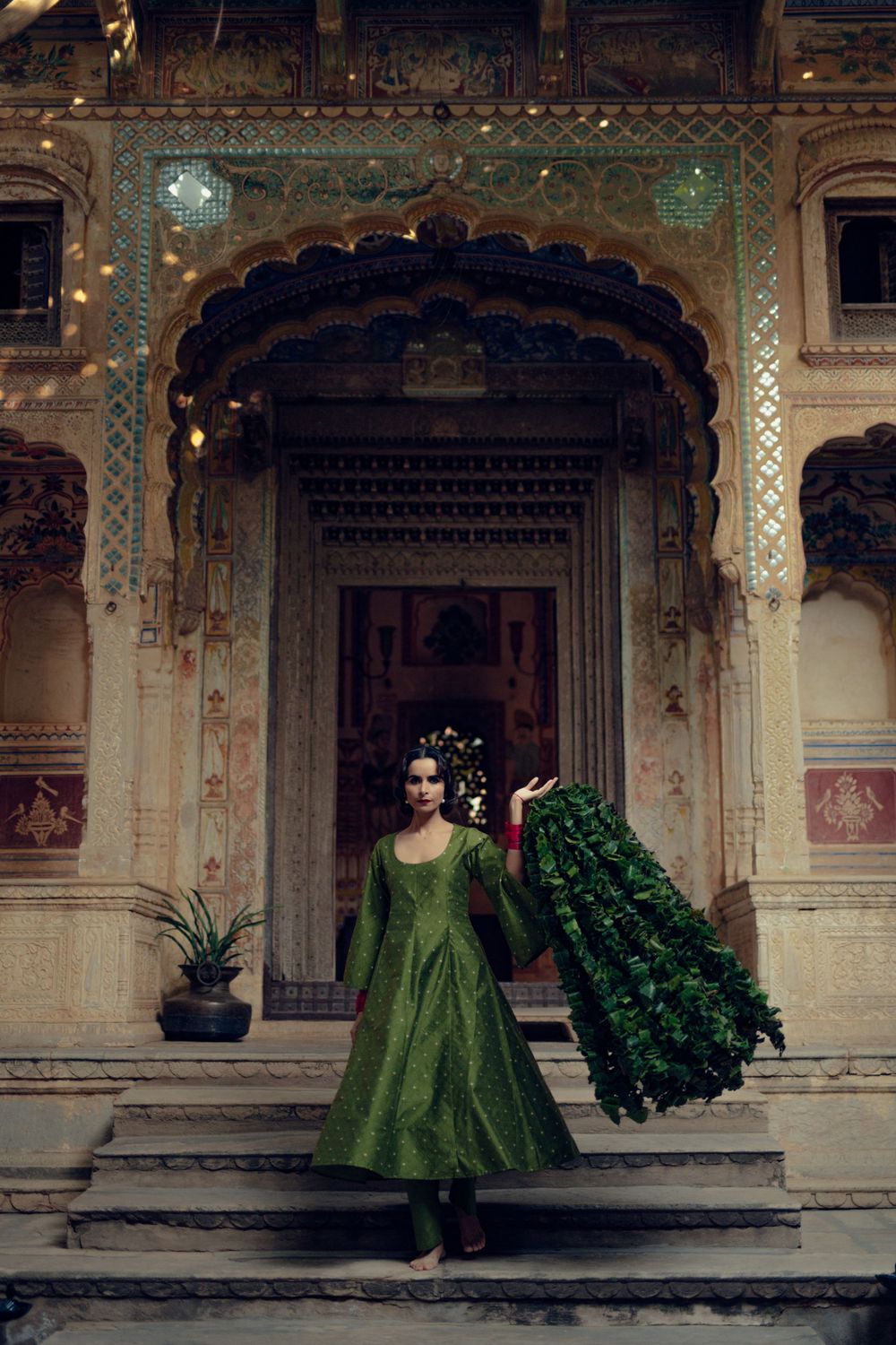 Green anarkali dress against ornate Indian palace architecture with peacock feathers.