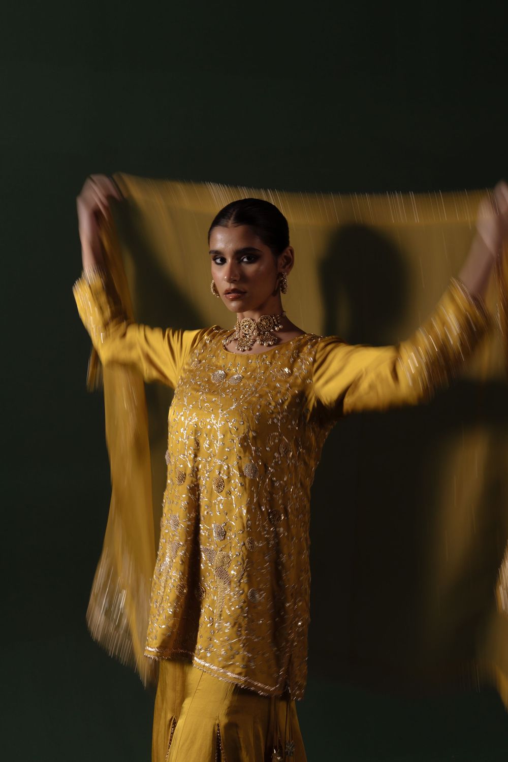 A figure wearing a shimmering golden embellished top performs an expressive dance movement against a dark background.