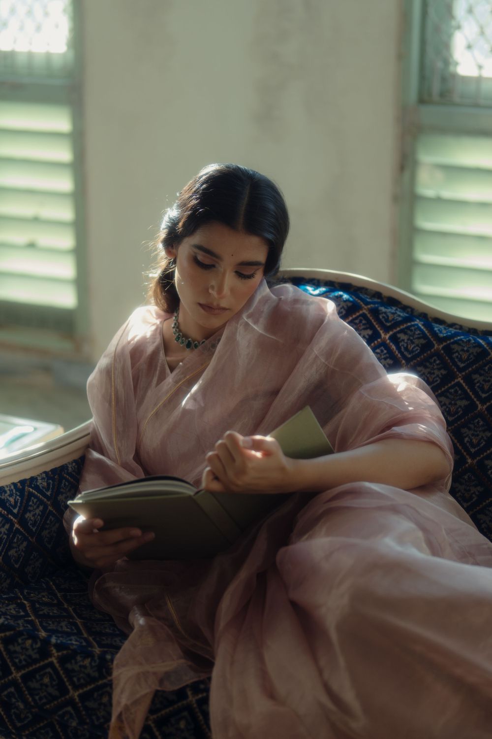 Peaceful scene of person in pink saree reading book on ornate blue couch.