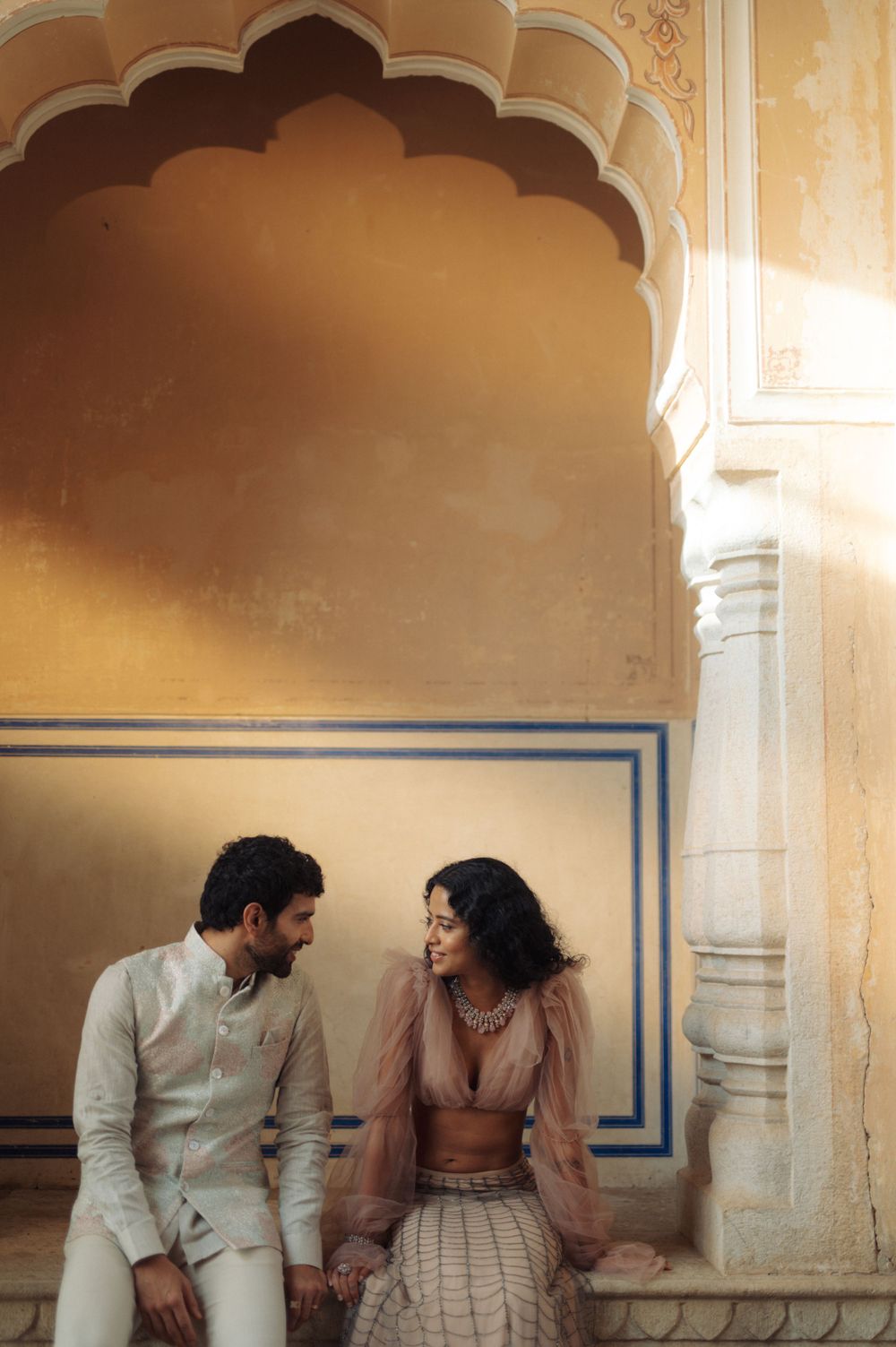 Romantic couple in light colored outfits sitting under ornate archway.