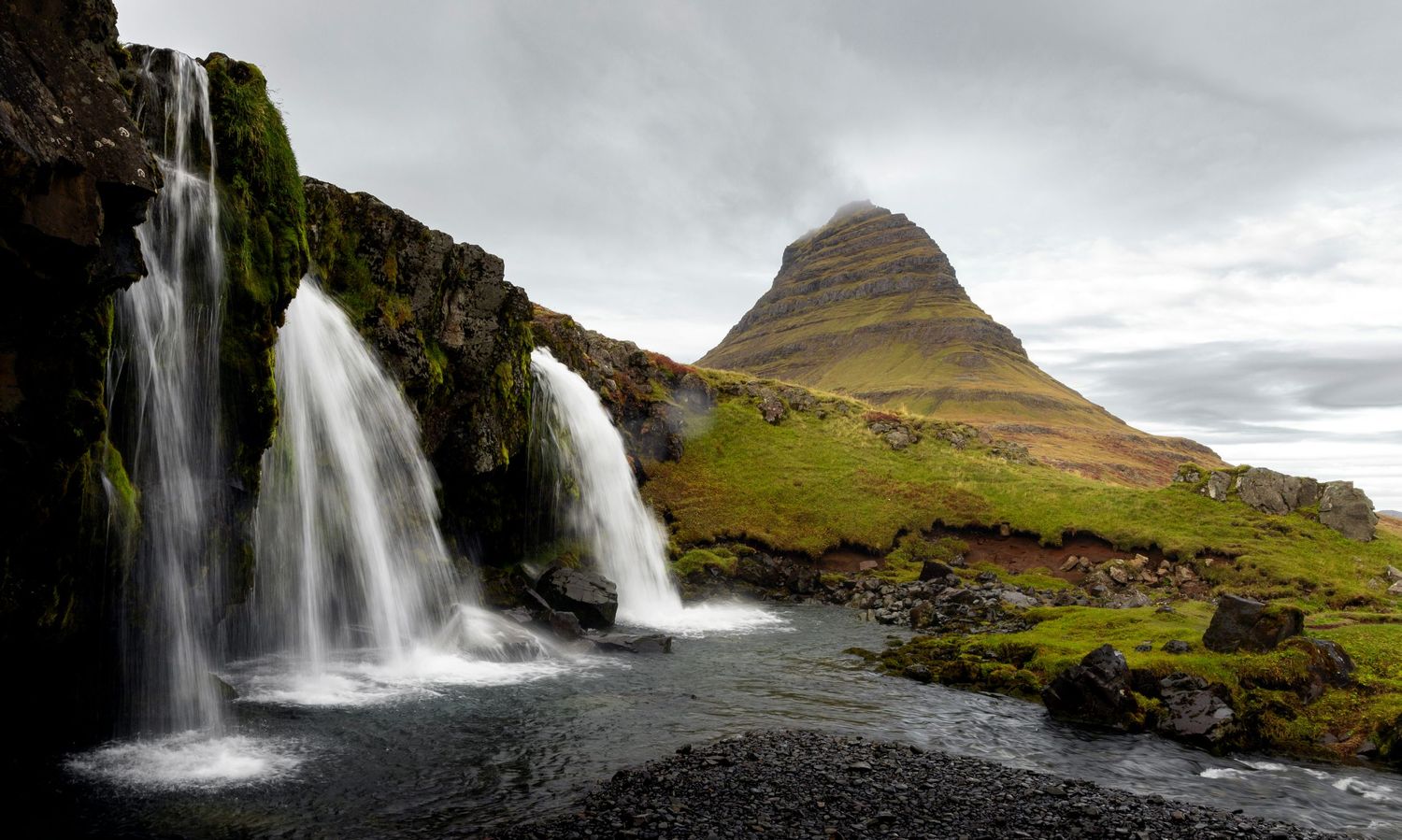 Kirkjufell mountain rises behind cascading waterfalls in Iceland's dramatic landscape under moody cloudy skies.