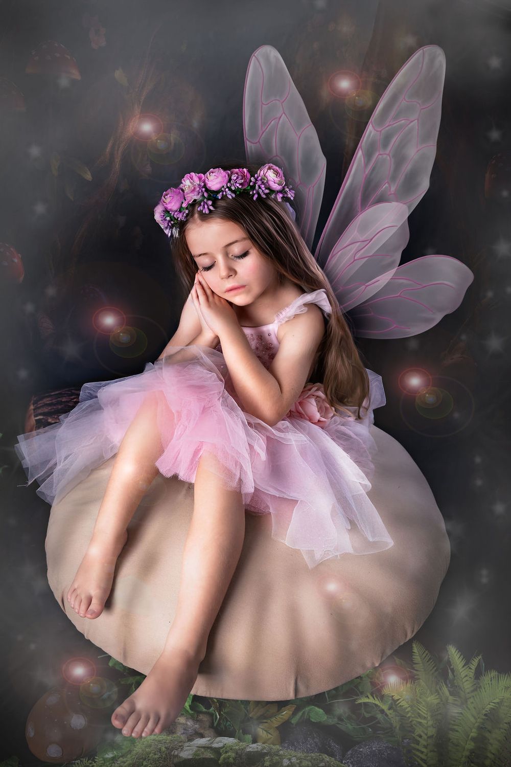 A young girl dressed as a fairy in a pink tutu dress with wings and flower crown sits on a mushroom.